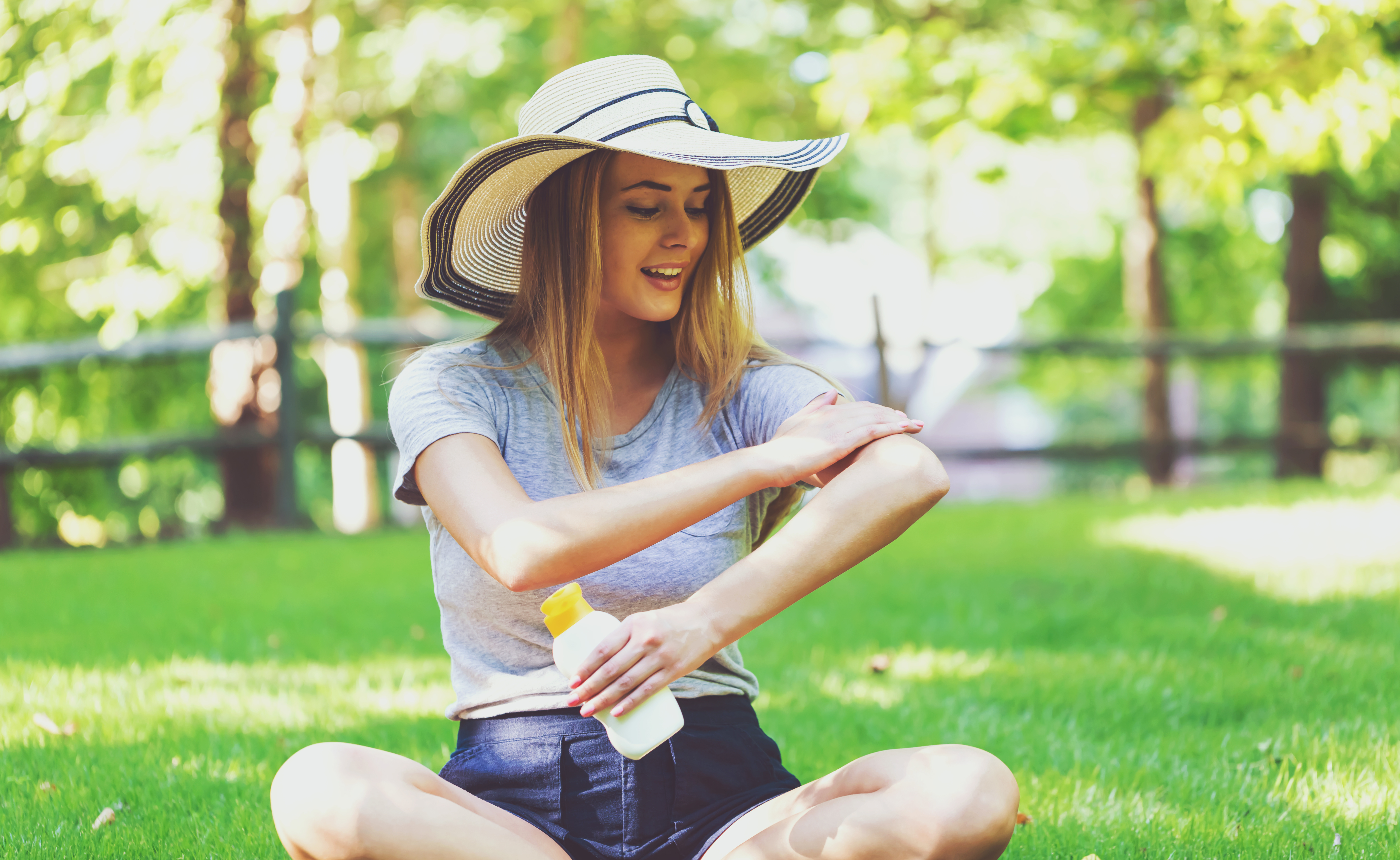 a young woman in a big hat applying sunscreen while sitting in the park on a nice day